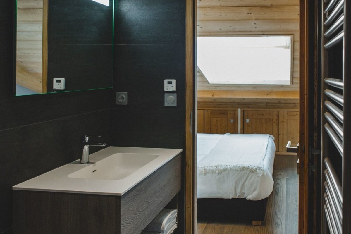 A modern, dark-tiled bathroom with a sink, looking through an open doorway into a warm, wood-paneled bedroom with a white bed.