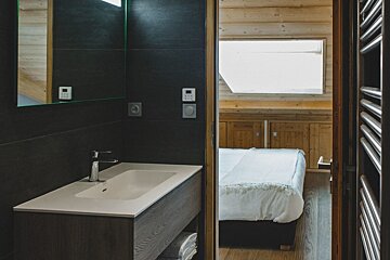A modern, dark-tiled bathroom with a sink, looking through an open doorway into a warm, wood-paneled bedroom with a white bed.