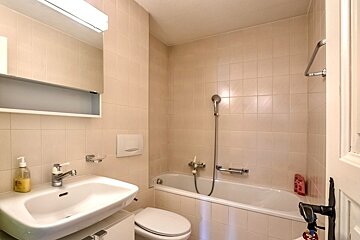 A light-tiled bathroom featuring a bathtub with an attached shower, a toilet, and a sink with a mirror cabinet above.