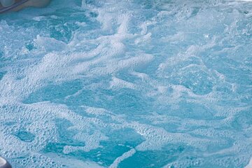 Vibrant blue water actively bubbling and foaming, filling the frame. It's likely a hot tub or jacuzzi with active jets, creating a textured, frothy surface.