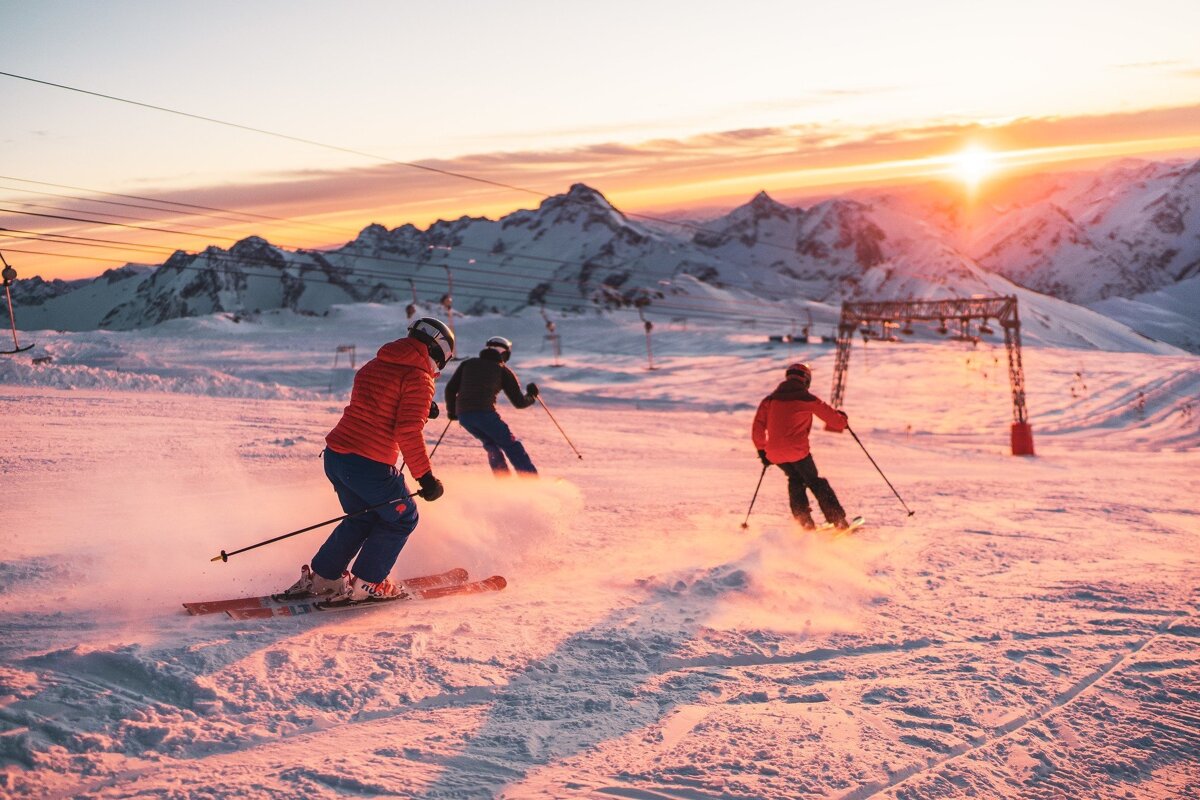 Three skiers descend a snowy mountain at a vibrant sunset/sunrise, casting an orange glow over the peaks and slopes as powder flies.