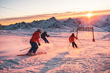 Three skiers descend a snowy mountain at a vibrant sunset/sunrise, casting an orange glow over the peaks and slopes as powder flies.