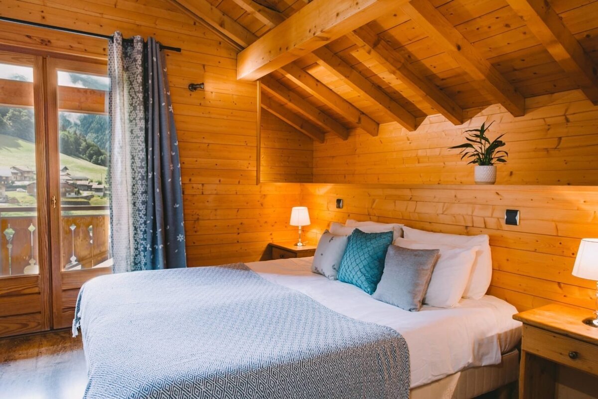 A cozy, rustic bedroom with warm wood paneling, a bed with blue accents, and a balcony view of mountains and a village.