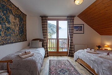 A cozy twin bedroom featuring a large tapestry, plaid curtains, and a balcony with a misty mountain view. A slanted wooden ceiling adds warmth.