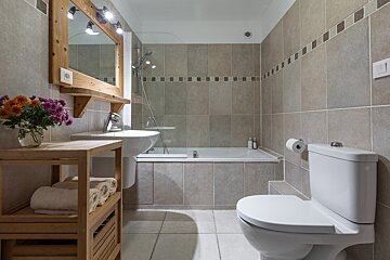 A cozy, tiled bathroom with a tub/shower, toilet, sink, and wooden mirror/shelving. Flowers and neatly folded towels add a homey touch.