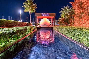 A vibrant night view of Bâoli: a reflecting pool leads to a glowing sphere under a sign, flanked by illuminated hedges and palm trees, with marina lights beyond.