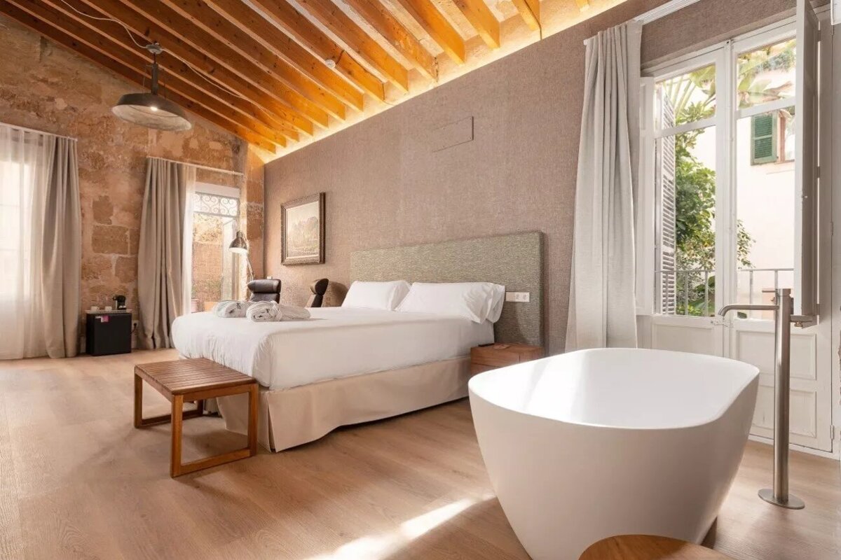 A chic bedroom blending rustic stone walls and exposed beams with a modern bed and a freestanding bathtub on a light wood floor.