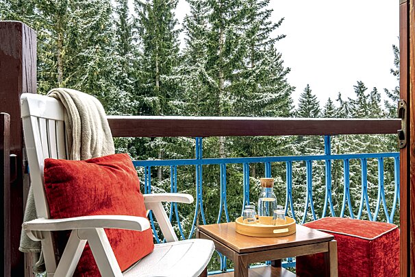 A chair with a red pillow sits on a balcony overlooking a snowy forest