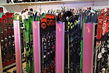 A ski shop with numerous colorful skis standing upright in rows, separated by pink dividers. Ski helmets are visible on shelves above.