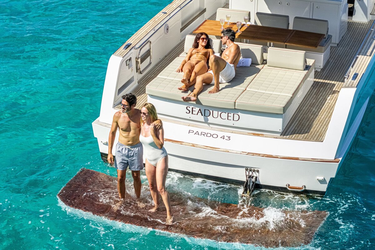 Two couples enjoying a luxury yacht in turquoise water, one lounging on deck and the other standing on the swim platform.