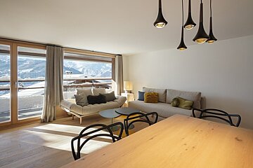 Bright modern living room with large windows overlooking snowy mountains. Features sofas, a dining table, and stylish pendant lights.