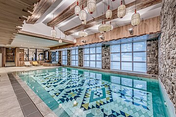 A rustic indoor pool featuring a vibrant geometric tile pattern, wooden beams, stone walls, and unique hanging lanterns, with a gym visible in the background.