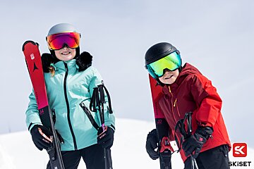 Two kids in ski gear, including helmets and goggles, smile while holding skis against a snowy background.
