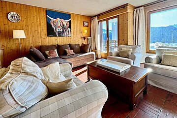 A cozy, wood-paneled living room with a colorful Highland cow painting, brown and plaid sofas, and large windows revealing a sunny mountain view.