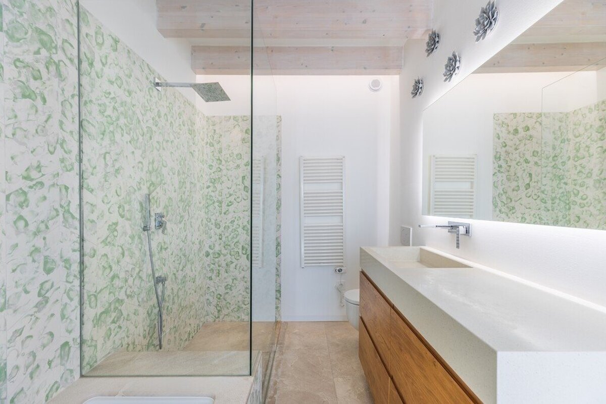 Modern bathroom with a glass shower featuring green patterned tiles, a long wooden vanity, and a large illuminated mirror.