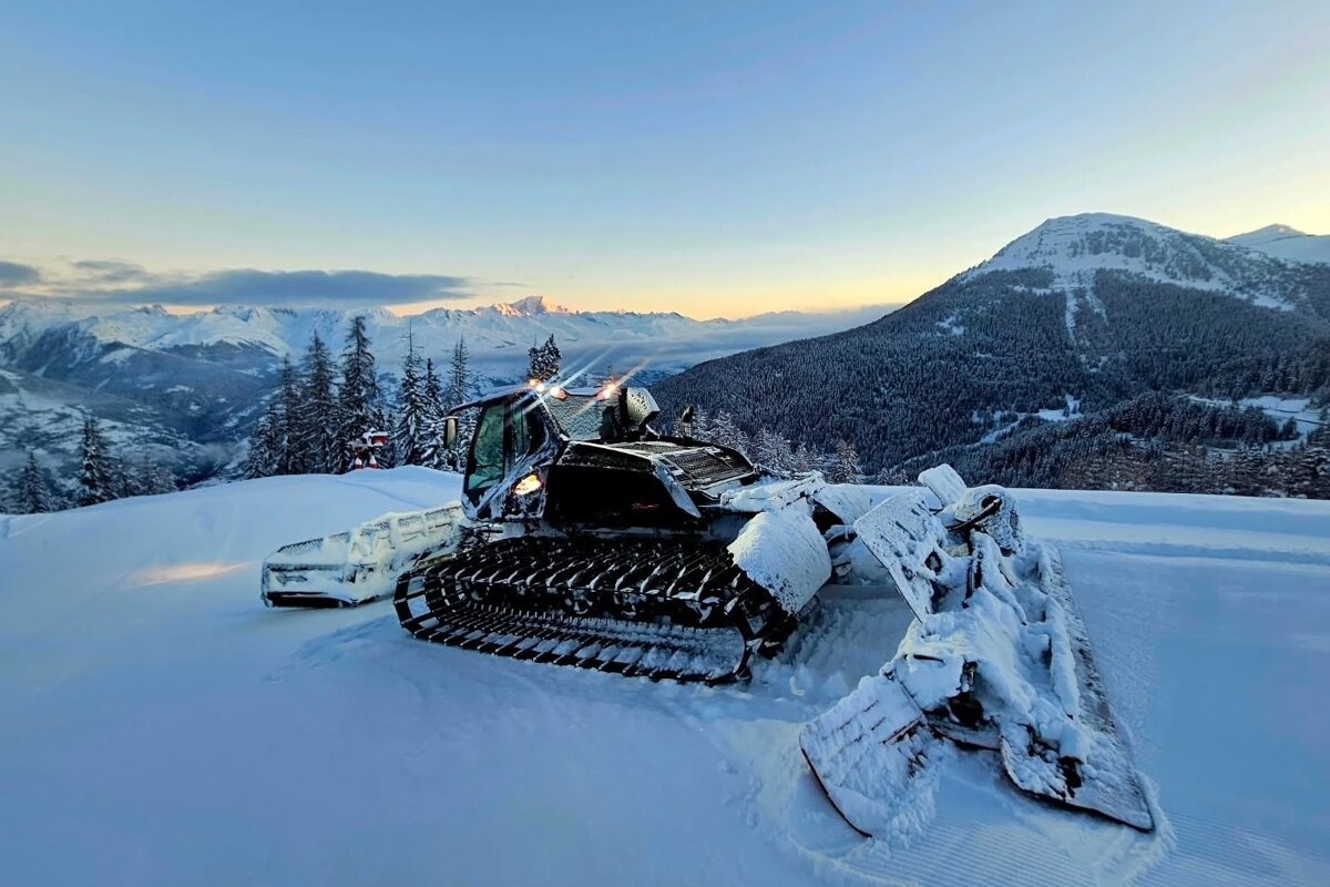 A snow groomer rests on a snowy piste with stunning mountain views, frosted trees, and a soft, colorful sky at dawn or dusk.