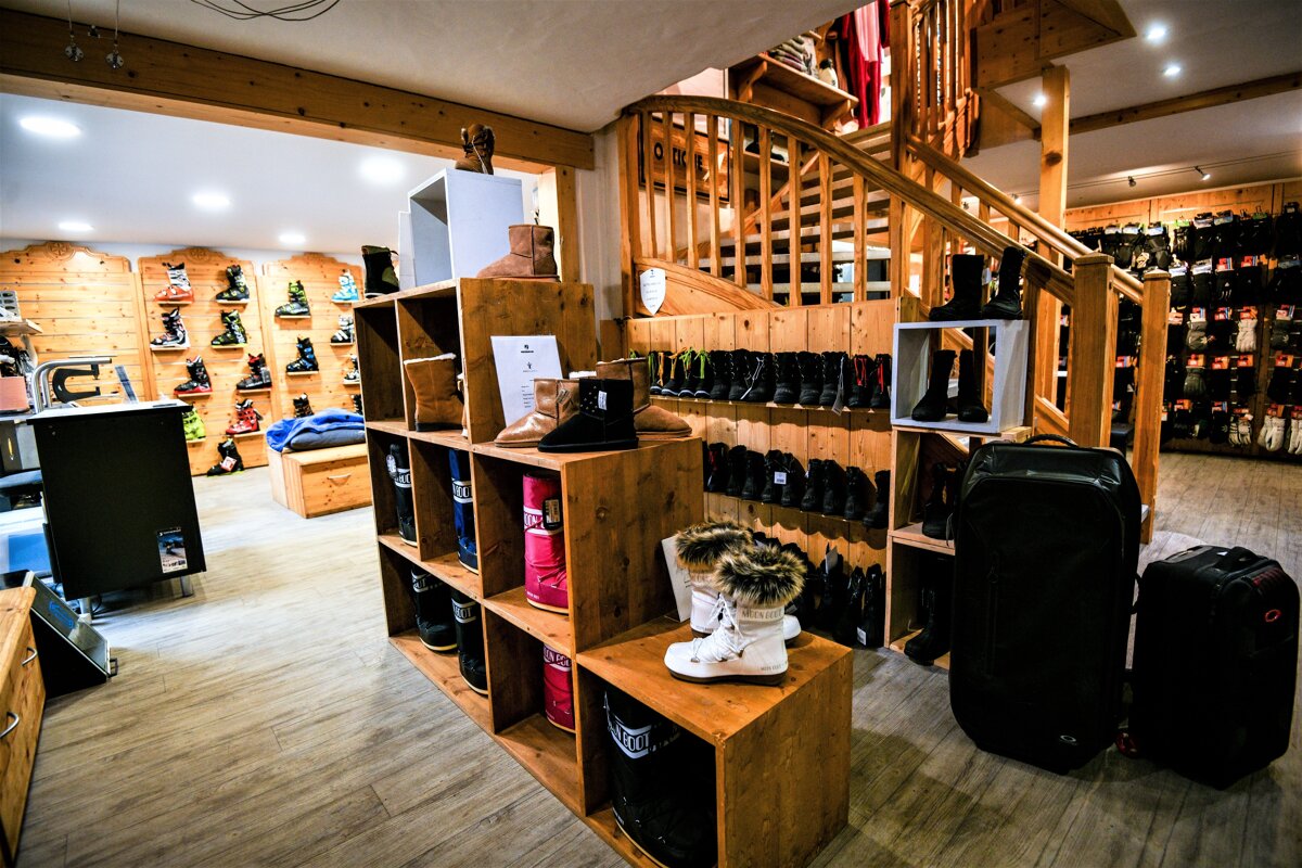 A rustic ski and winter boot shop with wooden shelves, walls, and a prominent staircase. Various colorful boots and outdoor gear are neatly displayed throughout.