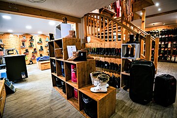 A rustic ski and winter boot shop with wooden shelves, walls, and a prominent staircase. Various colorful boots and outdoor gear are neatly displayed throughout.