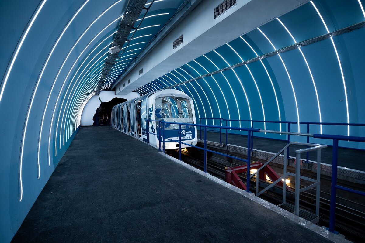 A modern, blue-hued train station with a white and blue train at the platform. Curved white light strips illuminate the tunnel's walls.