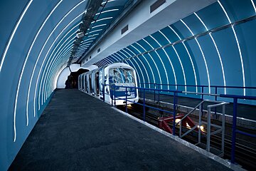 A modern, blue-hued train station with a white and blue train at the platform. Curved white light strips illuminate the tunnel's walls.