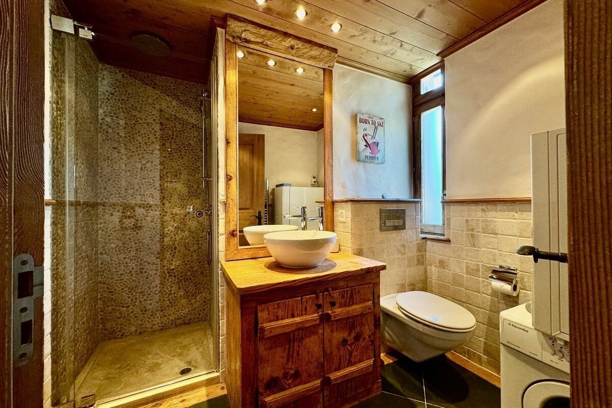 Rustic bathroom with wooden ceiling, vanity, and mirror. Features a pebble-tiled shower, white vessel sink, toilet, and washing machine.