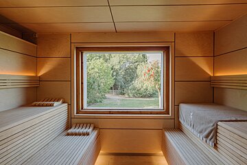 A contemporary wooden sauna features ambient lighting, slatted benches, and a wide window overlooking a vibrant green garden.