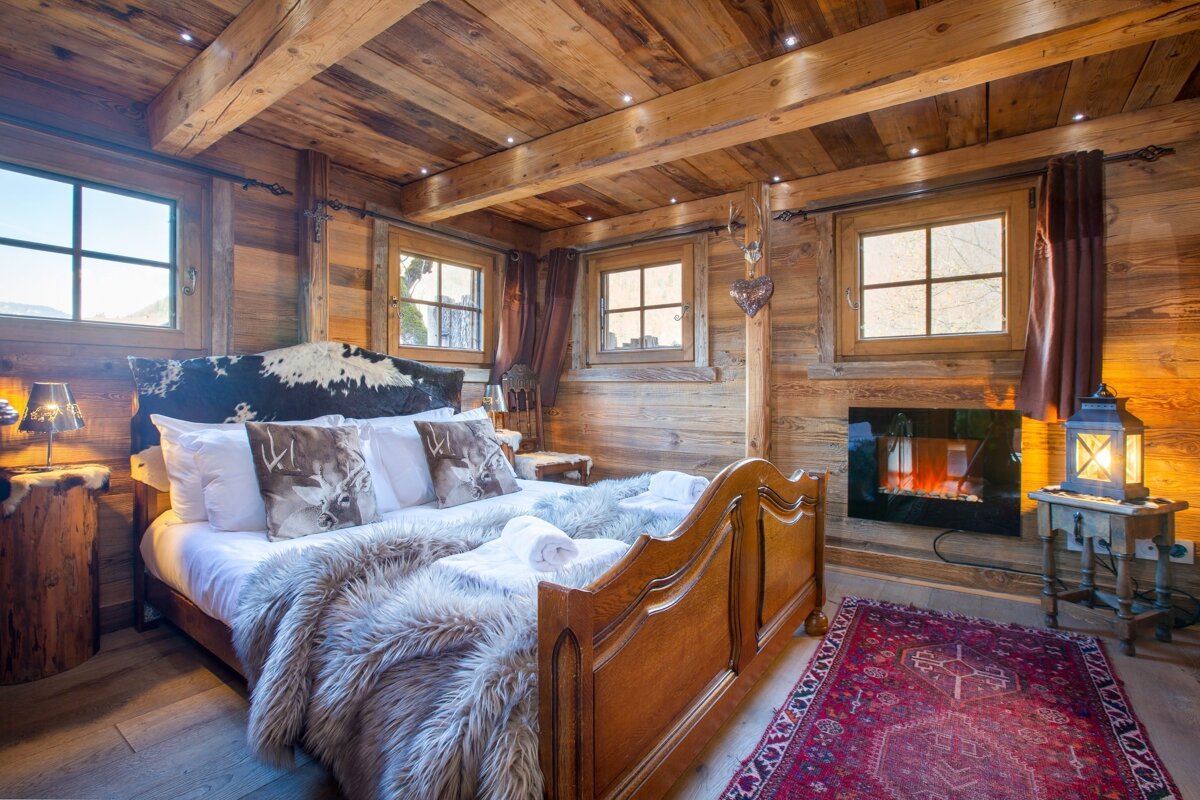Cozy, rustic wooden chalet bedroom with a large bed, fur throws, cowhide headboard, and an electric fireplace, illuminated by soft lighting.