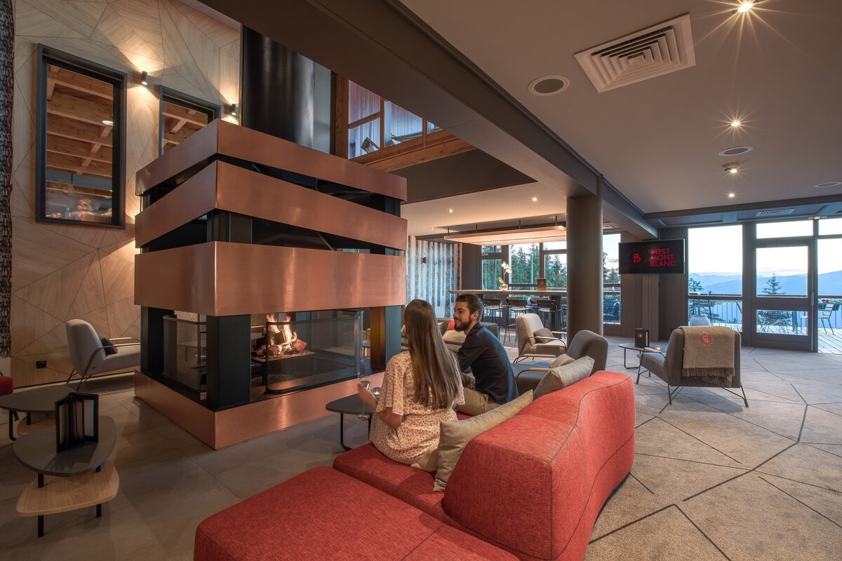 A couple relaxes on a red sofa by a modern copper fireplace in a stylish lounge with large windows offering mountain views, creating a cozy ambiance.