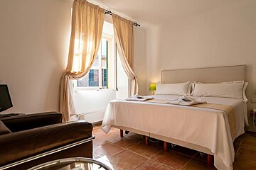 A cozy, brightly lit bedroom with a white bed, beige curtains on a window, brown tiled floor, a leather armchair, and a TV.