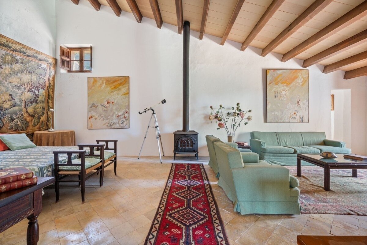 Spacious room with exposed beam ceiling, wood-burning stove, light green sofas, terracotta floor, tapestry, and telescope, creating a rustic yet cozy feel.
