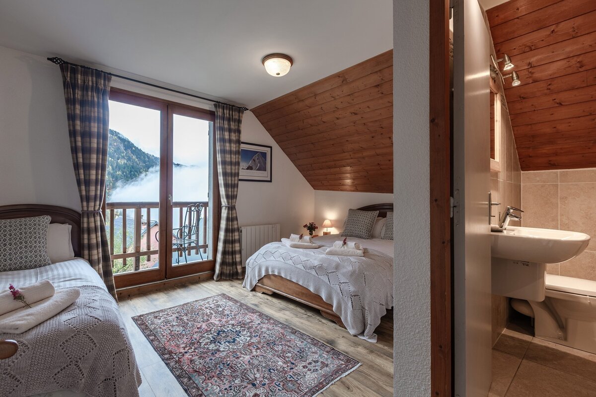 Cozy, rustic bedroom featuring a double and single bed, balcony with a foggy mountain view, and an en-suite bathroom. Wood-paneled ceiling adds warmth.