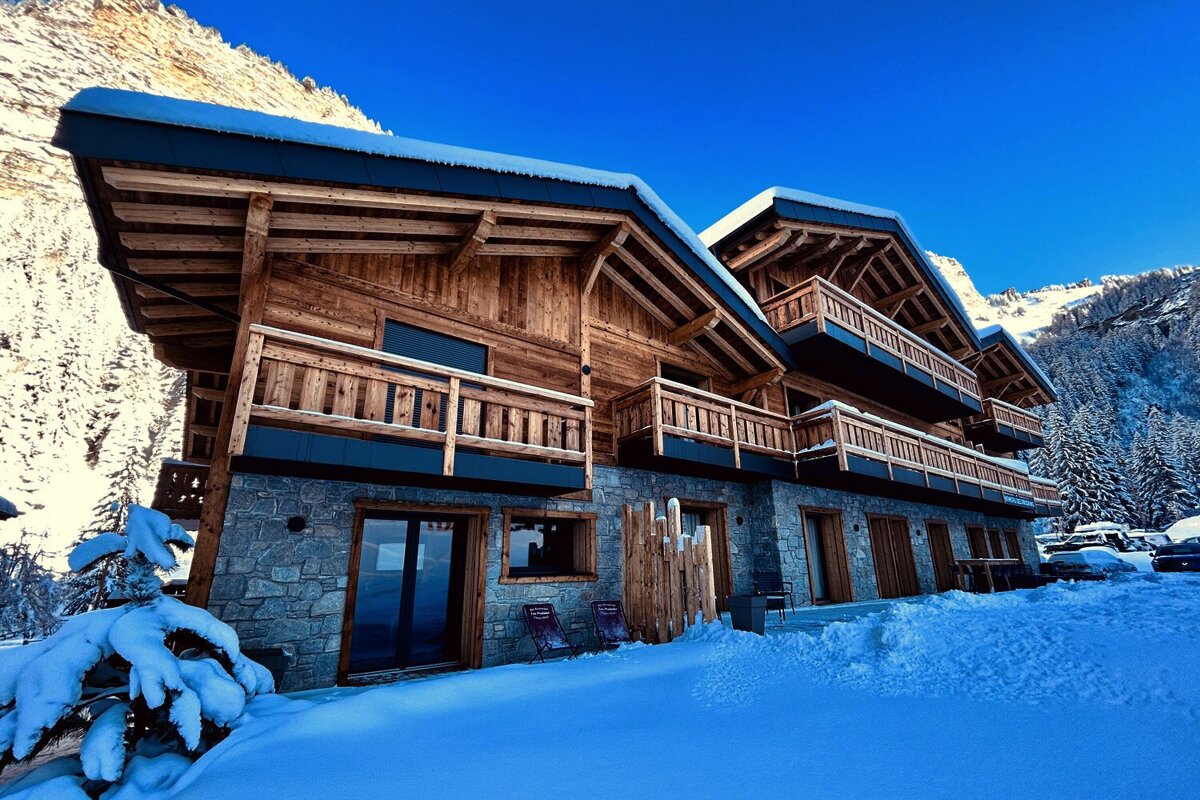 A grand wooden and stone chalet with balconies is surrounded by deep snow and majestic mountains under a bright blue sky. A beautiful winter scene.
