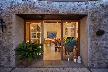 Rustic stone doorway frames glass sliding doors opening to a bright, inviting room with plants, wooden furniture, and ambient lighting.