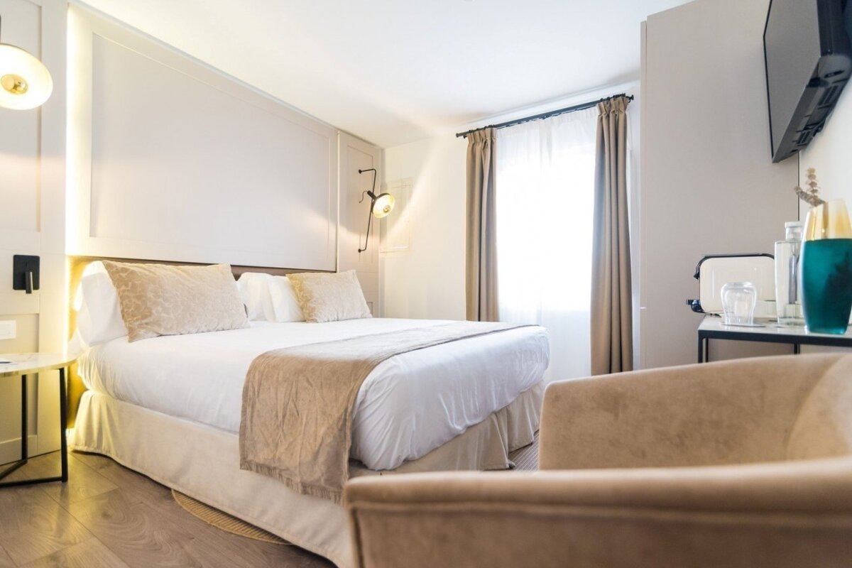 A bright, modern bedroom features a large bed with neutral bedding, a light-filled window, and an inviting armchair in the foreground.