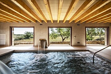 An indoor spa with a bubbling pool and waterfall under a beamed ceiling. Large windows frame a sunlit natural landscape with trees.