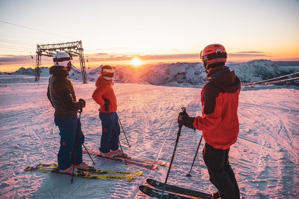 Three skiers admire a stunning sunset over snow-covered mountains from a ski slope, with distant peaks and ski lift structures visible.