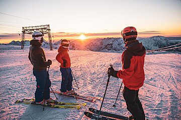 Three skiers admire a stunning sunset over snow-covered mountains from a ski slope, with distant peaks and ski lift structures visible.