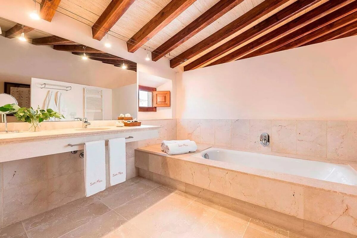 Elegant marble bathroom with a deep tub, double vanity, white towels, and a high ceiling with exposed wooden beams and track lighting.