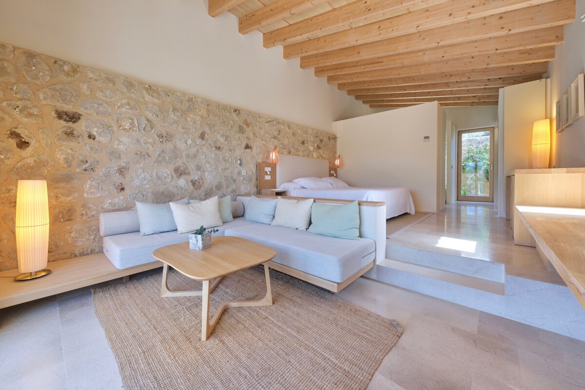 A bright, modern room featuring a stone wall, wooden beams, a cozy seating area, and a raised bedroom with a bed. Natural textures and minimalist design.