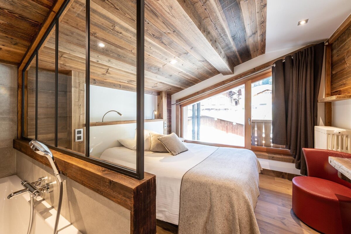 A cozy, rustic modern bedroom featuring a wood-paneled ceiling, large window, comfortable bed, and an integrated bathroom with a glass partition.