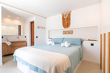 A bright, modern bedroom with a large bed, blue linens, and a macrame wall hanging. An open-concept bathroom with a wooden vanity is visible.