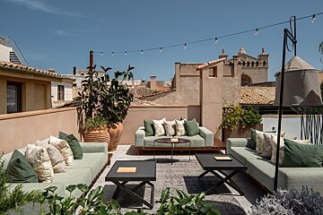 Chic rooftop lounge with green sofas, string lights, and lush plants, offering views of a Mediterranean town under a blue sky.