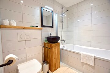 A bright, modern bathroom with white tiled walls, a toilet, a unique black bowl sink on a wooden stand, a bathtub with a shower, and a mirror.
