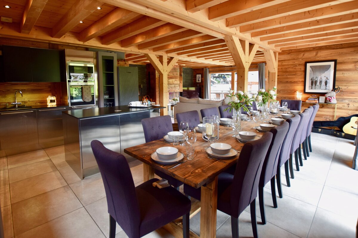 Spacious open-plan chalet kitchen and dining area. Features a large wooden table with purple chairs, a sleek stainless steel kitchen, and warm wood beams.