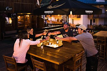 Four people toast with drinks around a glowing fire pit on a cozy outdoor patio at night, enjoying a social gathering.