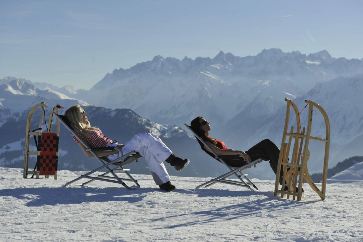 Two women sit in lawn chairs next to sleds in the snow