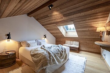 A cozy attic bedroom features slanted wooden ceilings and walls, a skylight, and a plush white bed with a fluffy blanket, creating a warm retreat.