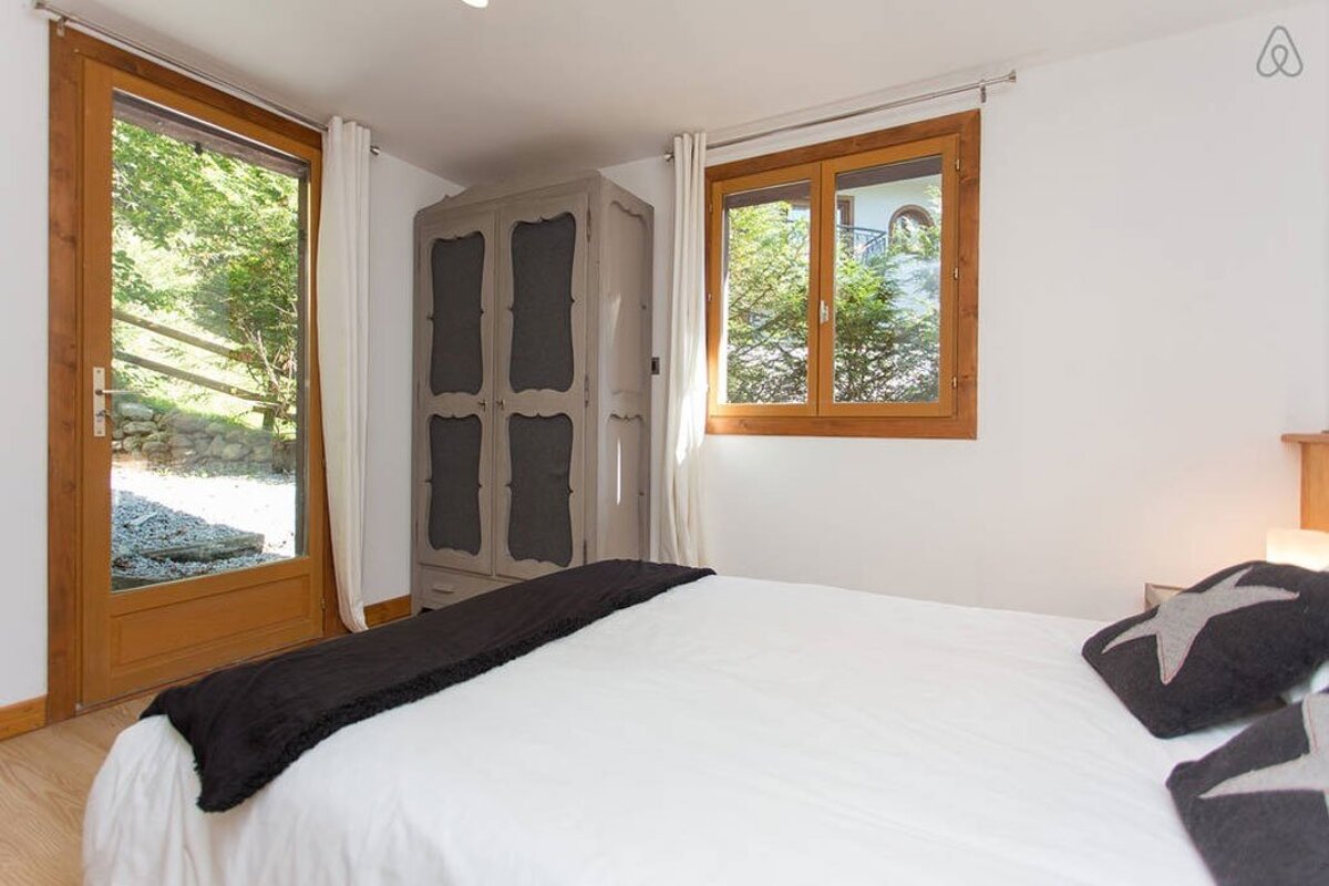 A serene bedroom features a white bed with dark pillows, a rustic grey wardrobe, and wooden-framed windows and a glass door opening to green nature.