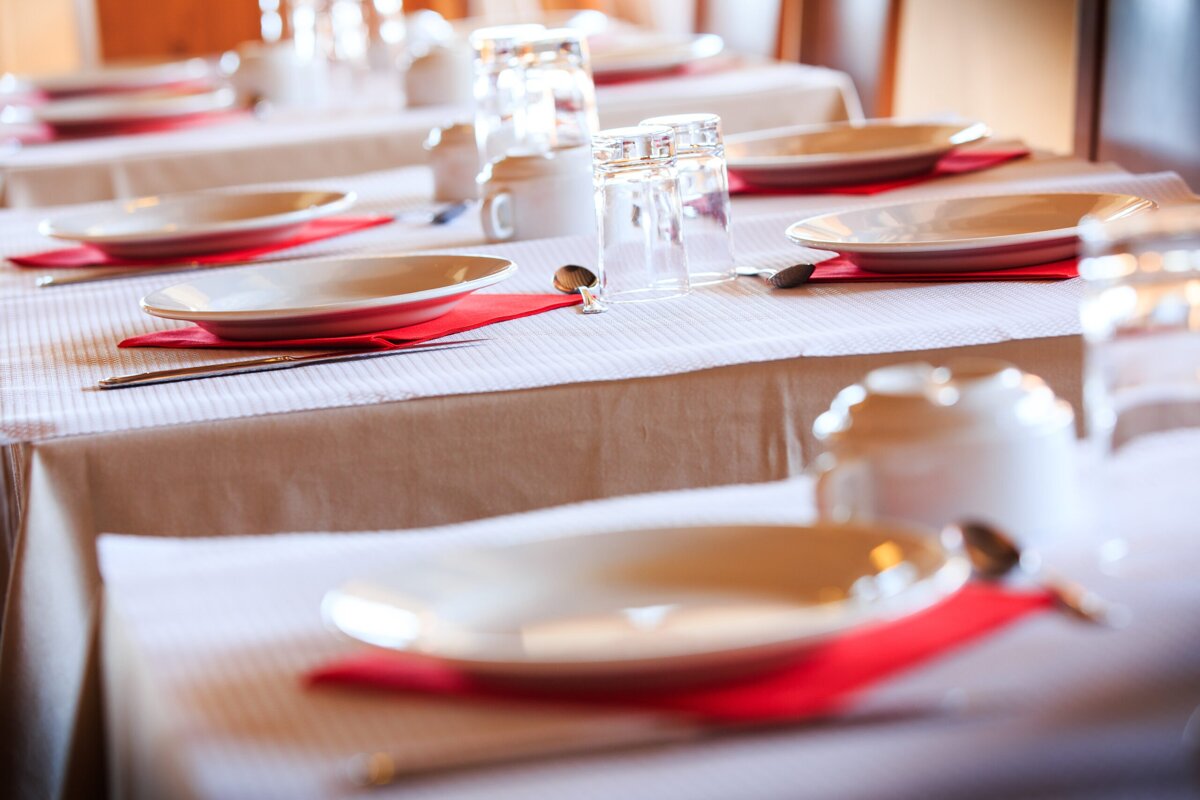 Multiple tables are set for dining with white tablecloths, plates on red napkins, glasses, and cutlery, ready for a meal.