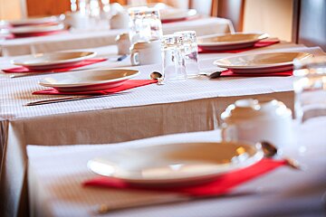Multiple tables are set for dining with white tablecloths, plates on red napkins, glasses, and cutlery, ready for a meal.